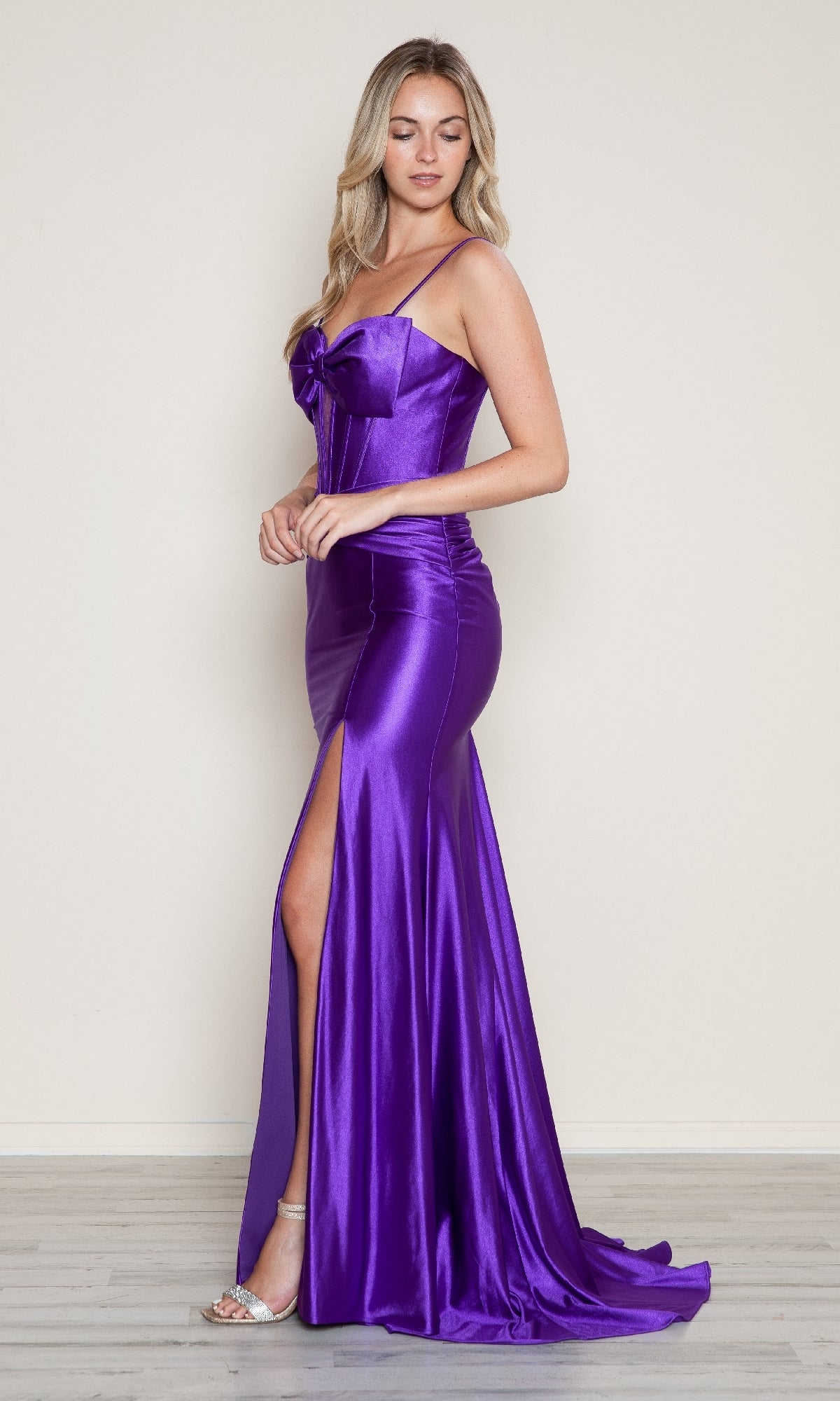A woman in a purple Front-Bow Long Cut-Out Prom Dress (Poly USA 9644) with spaghetti straps, a side slit, and bow bodice detail poses against a plain background, highlighting the gown’s elegant corset style.